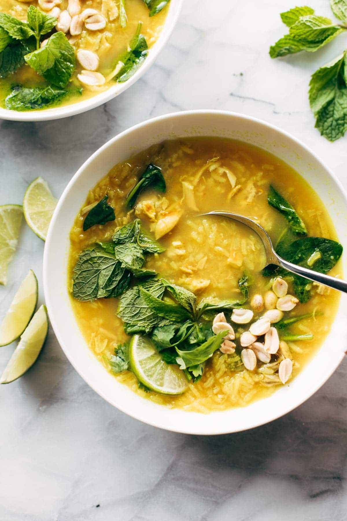 Chicken and rice soup in a bowl with a spoon. There are fresh herbs on top and lime wedges on the side. 