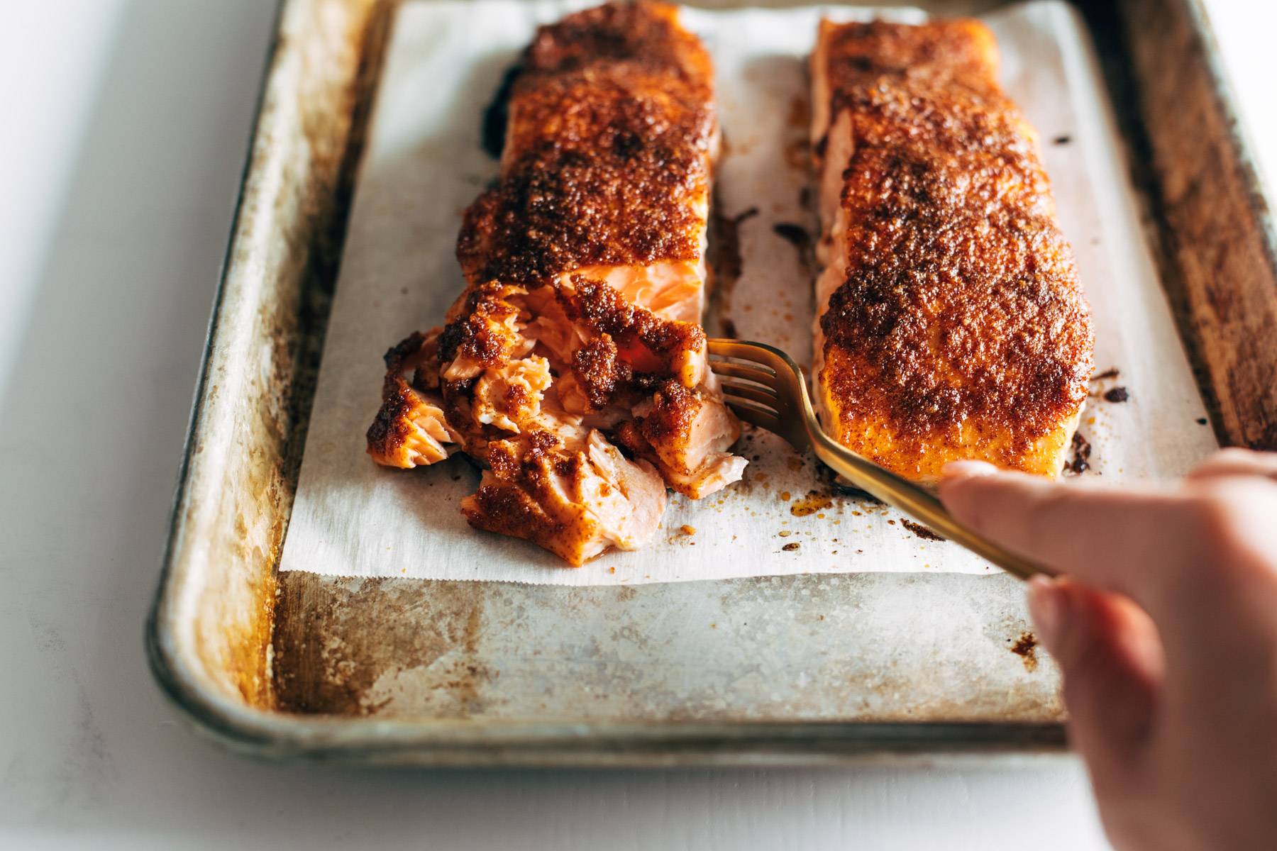 White hand flaking off a piece of salmon on a baking sheet